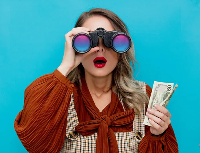 A woman with long hair looks through binoculars with purple lenses, holding a stack of cash in her left hand. She is wearing a brown blouse and patterned top against a solid blue background. Her lips are red, and her expression is surprised.