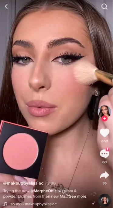 Woman applying blush with a brush, while holding a compact with pink blush. She has long hair, winged eyeliner, and pink lipstick. There are icons and text indicating social media interactions on the screen.