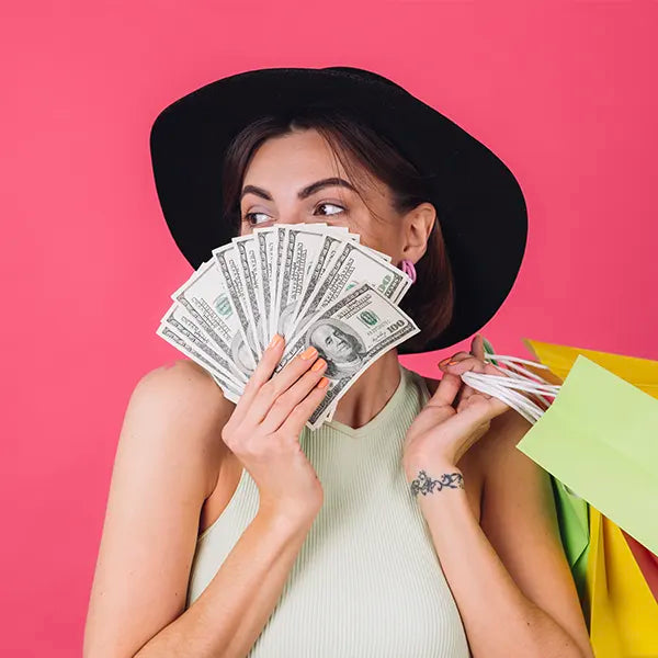 A woman in a black hat holds a fan of US dollar bills over her mouth. She has long hair and is wearing a sleeveless top. Her other hand holds colorful shopping bags. The background is pink.