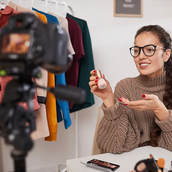 A woman wearing glasses and a brown sweater sits in front of a camera, holding a cosmetic product in one hand and gesturing with the other. Behind her, colorful clothes are hanging on a rack. A makeup palette is on the table.