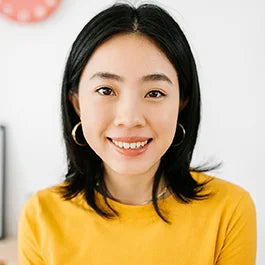 A person with medium-length black hair and hoop earrings is smiling and wearing a yellow top. The background is softly blurred.
