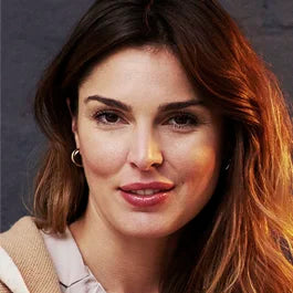 A woman with long brown hair and hoop earrings smiles softly. She is wearing a beige top and is positioned against a dark background. Her expression is friendly and relaxed.