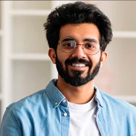 Man with a beard and glasses, wearing a blue shirt over a white top, smiles while standing indoors against a blurred, light-colored background.