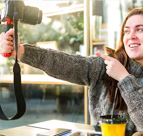 A person with long hair is smiling and pointing at a camera held in their outstretched hand. They are sitting indoors near a window, wearing a gray sweater. A table with a smartphone and a cup with a straw is in the foreground.