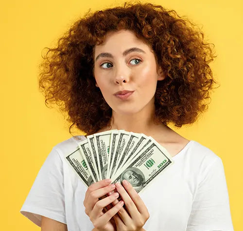 Woman with curly hair holding several hundred-dollar bills in her hand, looking to the side with a thoughtful expression. She is wearing a white shirt and stands against a bright yellow background.