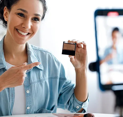 A smiling person in a blue denim shirt holds up a small makeup compact while pointing at it. They are seated indoors, with a smartphone on a tripod visible in the background, suggesting they may be recording a video.